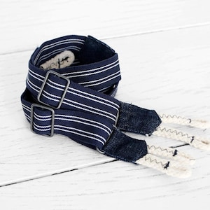 Peut inclure: Bretelles rayées bleu marine et blanches avec boucles métalliques. Les bretelles ont des détails en denim et des extrémités cousues en zigzag de couleur crème. L'accessoire est présenté sur une surface en bois blanc.