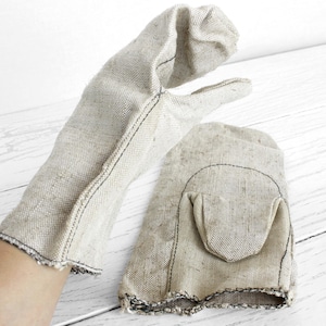 May include: Two beige oven gloves made from a textured fabric. One is on a hand, the other is lying flat. Both have black stitching and a pocket-like design on the palm. The background is a white wooden surface.