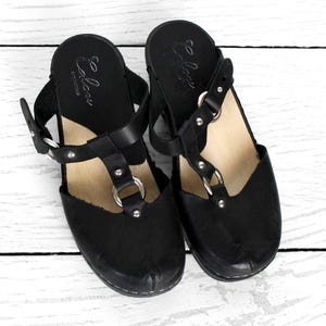 Pode incluir: Um par de tamancos de couro preto com design de tira em T. Os tamancos têm sola de madeira e detalhes em metal prateado. A marca "Elow" está impressa na palmilha. Os sapatos estão sobre um fundo de madeira branca.