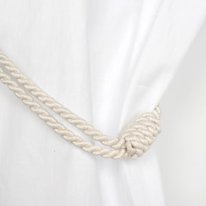 以下が含まれることがあります： クリーム色のロープカーテンタッセルは、端で結ばれ、白いカーテンに掛けられています。ロープはねじれたストランドでできており、質感のある外観を作り出しています。背景は無地の白い生地です。