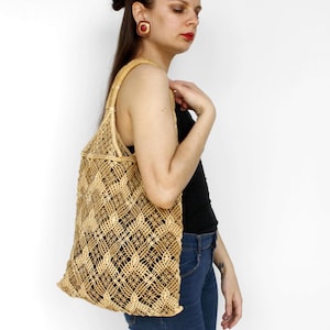 Könnte beinhalten: Eine beigefarbene, gewebte Tragetasche mit einem Rautenmuster. Die Tasche hat eine rechteckige Form und einen Griff aus dem gleichen Material. Die Tasche wird über der Schulter getragen. Der Hintergrund ist weiß.