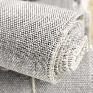 Può includere: Primo piano di un tessuto grigio arrotolato. Il tessuto presenta una trama tessuta e bordi sfilacciati. Un filo naturale avvolge il rotolo. Il materiale sembra essere lino o un tessuto simile, adatto a vari progetti artigianali.
