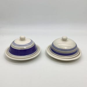 May include: Two ceramic butter dishes with lids. The dishes are white with blue and black stripes. Each lid has a small, round knob on top. The dishes are on a white surface.