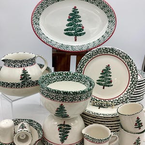 May include: A collection of white ceramic Christmas dinnerware featuring a green Christmas tree design. The set includes an oval platter, bowls, plates, mugs, a creamer, and a salt and pepper shaker set. Each piece has a green and red patterned border.