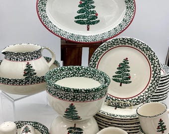 Op de afbeelding: Een verzameling wit keramisch kerstdinergoed met een groen kerstboomontwerp. De set bevat een ovale schaal, kommen, borden, mokken, een melkkan en een zout- en peperstel. Elk stuk heeft een groen en rood patroon.