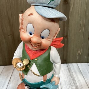 May include: A ceramic figurine of a cartoon character wearing a blue cap with a green four-leaf clover, a red bandana, a green vest, and blue pants. The character is holding a gold pocket watch and a small squirrel figurine.