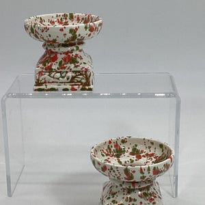 May include: Two white ceramic candle holders with red and green splatter paint. The holders have a square base and a round top.