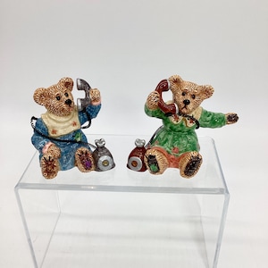 May include: Two ceramic bear figurines, each holding a telephone receiver. One bear wears a blue outfit, the other a green outfit. Both have small sacks at their feet. The figurines are on a clear acrylic display.