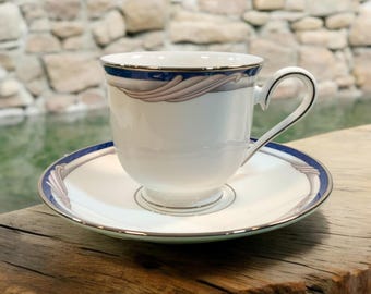 May include: A white teacup and saucer set with a blue and gray decorative border. The teacup has a curved handle and sits on a matching saucer. The set is placed on a wooden surface.