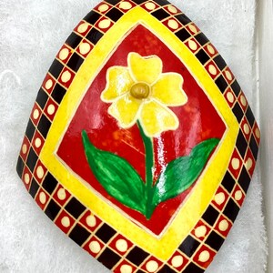 May include: A yellow flower with green leaves painted on a red background. The background is surrounded by a yellow border and a black and white checkered border.