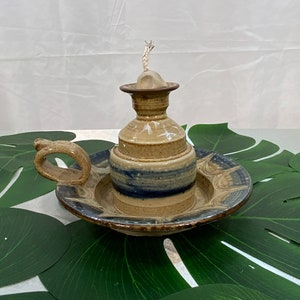 May include: A handmade ceramic oil lamp with a beige and blue color scheme. The lamp features a round base with a handle, a central reservoir, and a wick. The lamp sits on a matching plate with a decorative rim. The overall design is rustic and artisanal.