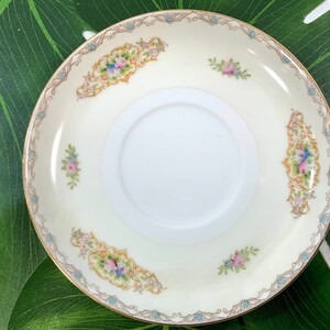 Op de afbeelding: Cr&egrave;mekleurige schotel met bloemen- en gouden rand. De schotel heeft een centrale witte cirkel en is versierd met roze, blauwe en gele bloempatronen. De rand heeft een gouden rand met een delicaat blauw en goud ontwerp.