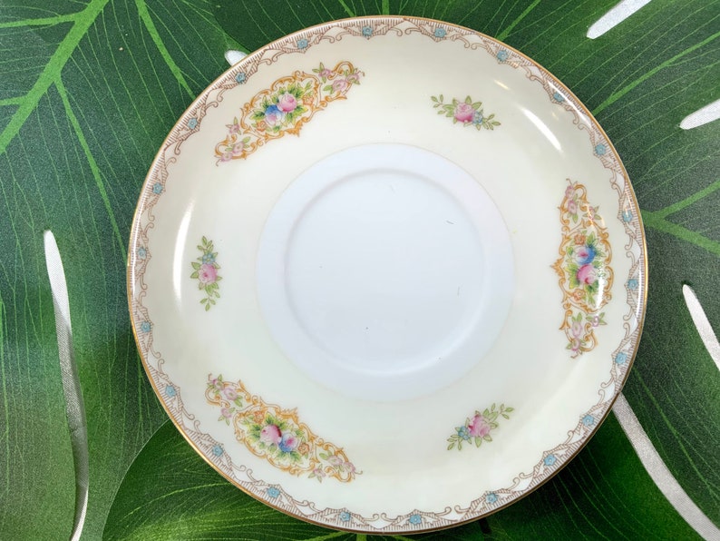 Op de afbeelding: Cr&egrave;mekleurige schotel met bloemen- en gouden rand. De schotel heeft een centrale witte cirkel en is versierd met roze, blauwe en gele bloempatronen. De rand heeft een gouden rand met een delicaat blauw en goud ontwerp.