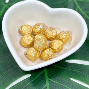 May include: A white heart-shaped dish filled with golden-wrapped chocolate candies. The candies are round and have a textured, foil-like wrapping. The dish is set against a green and white leaf-patterned background.