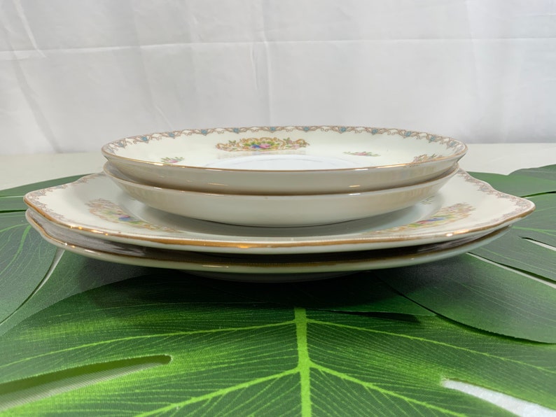 Op de afbeelding: Een stapel vintage porseleinen borden. De borden hebben een cr&egrave;mekleurige basis met gouden rand en een bloemenontwerp in het midden. Het bovenste bord is rond en het onderste bord is vierkant met geschulpte randen.