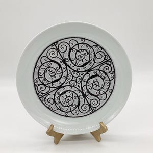 May include: White ceramic plate with a black and white design of a circular pattern of swirling lines. The plate is on a gold stand.