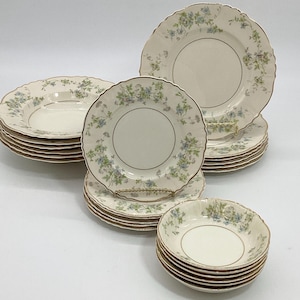 May include: A collection of vintage cream-colored china with a floral pattern in blue and green. The set includes dinner plates, salad plates, and bowls, all with gold rims and scalloped edges. A classic tableware set.