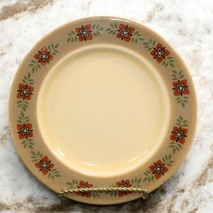 May include: A beige ceramic plate with a floral design. The rim features a repeating pattern of red and orange flowers with green leaves. The plate has a cream-colored center and is displayed on a gold-colored metal stand.