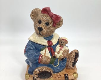 Puede incluir: Jarrón de galletas de cerámica con forma de oso de peluche que lleva un traje de marinero azul y un lazo rojo. El oso sostiene un oso de peluche más pequeño y un velero. El jarrón de galletas está sentado sobre una maleta marrón con un calcetín a rayas azules y rojas que sobresale.