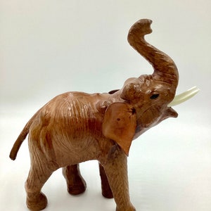 May include: A brown elephant figurine with its trunk raised and ivory-colored tusks. The elephant has large ears and is standing on four legs. The figurine is made of a textured material, possibly leather or a similar material.