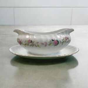 May include: A white porcelain gravy boat with a pink floral design and a matching saucer. The boat has a scalloped edge and a gold rim.
