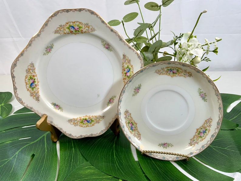 Op de afbeelding: Een witte porseleinen bord- en schotelset met gouden rand en bloemaccenten. Het bord is vierkant met geschulpte randen en de schotel is rond. Beide stukken hebben een decoratieve rand met vogels en bloemen.