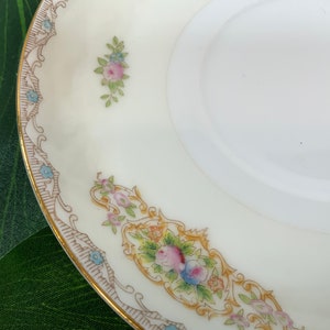 Op de afbeelding: Een witte porseleinen schotel met een gouden rand en bloemaccenten. De schotel heeft een decoratieve rand met een herhalend patroon en kleine clusters van roze en blauwe bloemen. Het ontwerp is delicaat en vintage-ge&iuml;nspireerd.