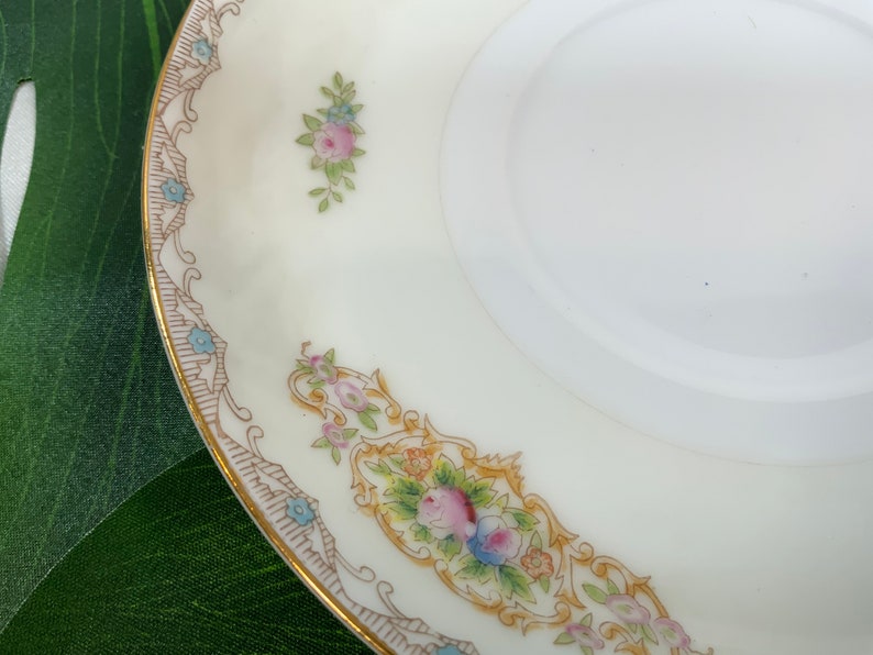 Op de afbeelding: Een witte porseleinen schotel met een gouden rand en bloemaccenten. De schotel heeft een decoratieve rand met een herhalend patroon en kleine clusters van roze en blauwe bloemen. Het ontwerp is delicaat en vintage-ge&iuml;nspireerd.