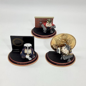 May include: Three decorative figurines featuring bulldog characters in military uniforms. One figure is in a sailor uniform, another is holding a gun in front of a brick wall, and the third is holding a gold coin. Each figurine is on a black base.