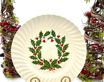 May include: A cream-colored ceramic plate with a scalloped, fluted edge and a central illustration of a green holly leaf wreath with bright red berries. The plate is supported by a gold-toned metal stand with two curved, decorative feet.