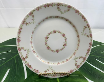 May include: White ceramic plate with a delicate floral design. The plate features a ring of pink flowers and green leaves around the rim and a smaller floral ring in the center. A gold-colored stand supports the plate.