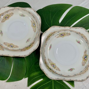 Op de afbeelding: Twee sets vintage porseleinen borden en schotels. De borden zijn vierkant met geschulpte randen en de schotels zijn rond. Elk stuk heeft een cr&egrave;mekleurige achtergrond met bloemen en gouden randen.