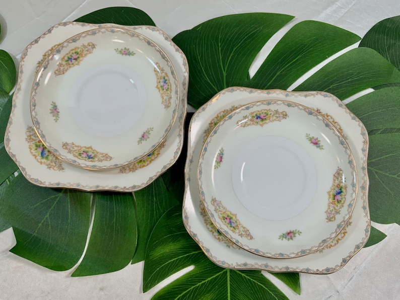 Op de afbeelding: Twee sets vintage porseleinen borden en schotels. De borden zijn vierkant met geschulpte randen en de schotels zijn rond. Elk stuk heeft een cr&egrave;mekleurige achtergrond met bloemen en gouden randen.