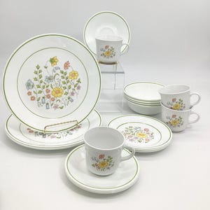 May include: A white floral dinnerware set with a green trim. The set includes plates, saucers, bowls, and cups. The floral design features yellow, blue, and orange flowers. The plates are approximately 10 inches in diameter.
