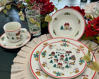 Op de afbeelding: Een wit keramieken servies met een feestelijke kerstscène van huizen, dennenbomen en figuren in een arrenslee. De onderdelen zijn voorzien van rode randen en een decoratie van gouden klokjes en hulstbladeren, inclusief een kopje en borden.