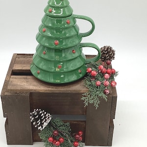 May include: A green ceramic Christmas tree-shaped mug with a gold star on top and red dot ornaments. The mug is on a wooden crate with pinecones and red berry sprigs, creating a festive holiday scene.