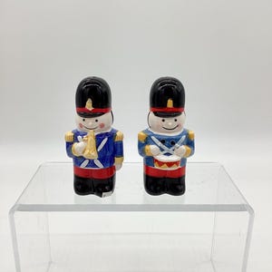 May include: Two ceramic toy soldier figurines. Each soldier wears a black hat, blue jacket, and red pants. One holds a gold trumpet, the other a drum. The figurines are on a clear, glass-like surface.