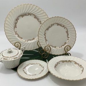 May include: A cream-colored dinnerware set with gold accents. The set includes plates, a bowl, and a covered sugar bowl. The plates have a spiral design and a gold trim. The sugar bowl has a floral design on the lid.