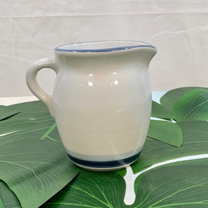 May include: A small, off-white ceramic pitcher with a curved handle. The pitcher has a dark blue band at the base and a thin blue line around the rim. The pitcher is likely used for serving liquids.