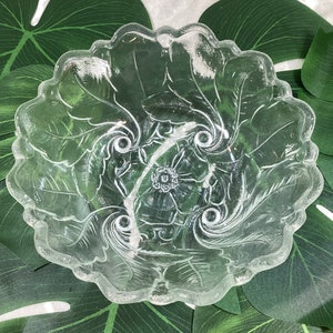 May include: Clear glass serving bowl with a floral and leaf design. The bowl has a scalloped edge and is resting on large green leaves. The bowl's interior features a central floral motif and swirling leaf patterns.