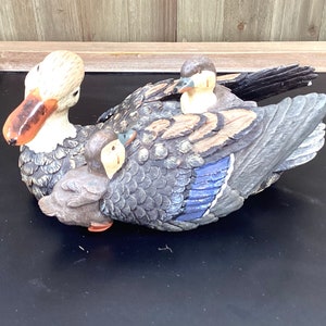 May include: A group of three carved wooden ducks. The ducks are painted in shades of brown, black, and blue. The largest duck has a white head and a brown beak. The ducks are arranged in a huddle, with the smaller ducks nestled under the larger duck.