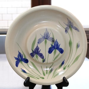 May include: A cream-colored ceramic serving bowl with a hand-painted iris design. The bowl features blue and purple irises with green stems and leaves. The bowl is displayed on a black stand.