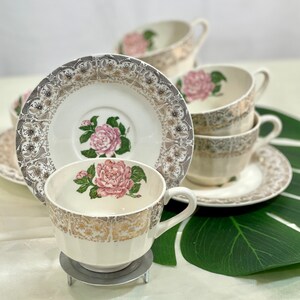 May include: A set of vintage teacups and saucers. The cups and saucers are cream-colored with a pink rose design in the center and gold trim. The saucers have a decorative border. The teacups are stacked on a white surface.