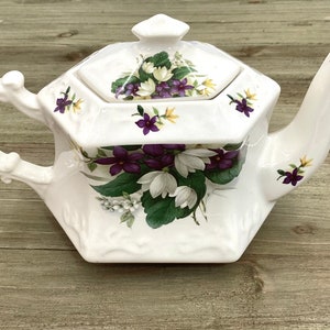 May include: White ceramic teapot with a hexagonal body and a lid. The teapot is decorated with a floral pattern of purple and white flowers and green leaves.