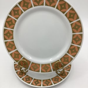 May include: Two white ceramic plates with a geometric design in orange and olive green. The plates are displayed on a gold-colored metal stand. The design features a repeating pattern of squares and diamond shapes around the plate's rim.