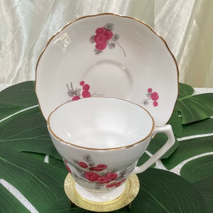 May include: White teacup and saucer set with a gold rim and a floral design featuring red berries. The teacup sits on a small, ornate gold stand. The saucer has a scalloped edge.