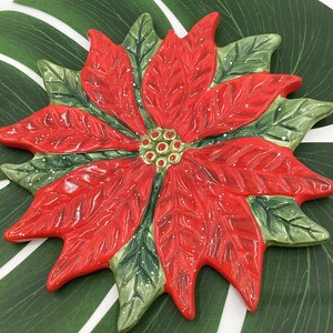 May include: A ceramic poinsettia flower with red petals and green leaves. The flower has a detailed design with white accents and a textured surface. The center of the flower is a cluster of small, round, green and gold details. The poinsettia is a festive holiday decoration.
