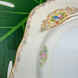 Op de afbeelding: Een cr&egrave;mekleurige, vierkante plaat met een gouden rand en bloemmotief. De plaat heeft een decoratieve rand met kleine blauwe bloemen en een centraal bloemarrangement met roze, blauwe en gele bloesems. De plaat heeft een geschulpte rand.