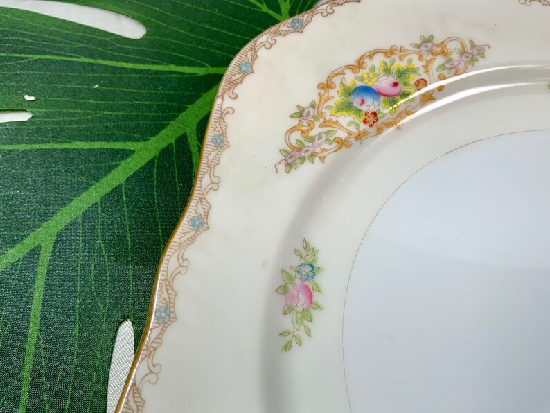 Op de afbeelding: Een cr&egrave;mekleurige, vierkante plaat met een gouden rand en bloemmotief. De plaat heeft een decoratieve rand met kleine blauwe bloemen en een centraal bloemarrangement met roze, blauwe en gele bloesems. De plaat heeft een geschulpte rand.