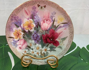 May include: A decorative porcelain plate with a gold rim and a floral design featuring pink, white, and yellow tulips, purple hyacinths, and a butterfly. The plate is on a gold stand.